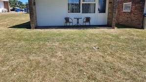 A single-story mobile home with a white exterior, a garage door, and two windows. There are two chairs outside the garage. - M & T Chalets , Detached Modern 2 bed Chalet, Sleeps 6 (Hemsby)