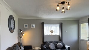 A modern living room with a sofa, a small side table, a wall clock, and a ceiling light fixture. - M & T Chalets , Detached Modern 2 bed Chalet, Sleeps 6 (Hemsby)