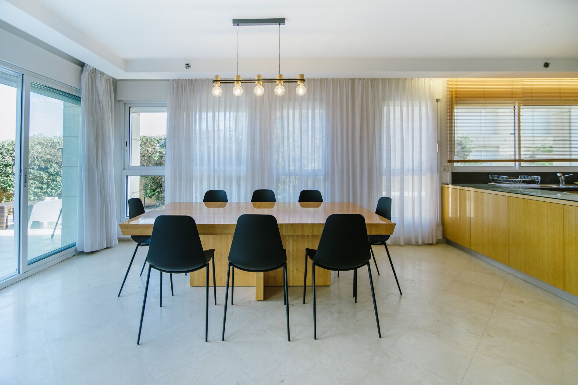 A modern dining area with a wooden table and black chairs, large windows with sheer curtains, and a kitchen area with wooden cabinets and a countertop.
