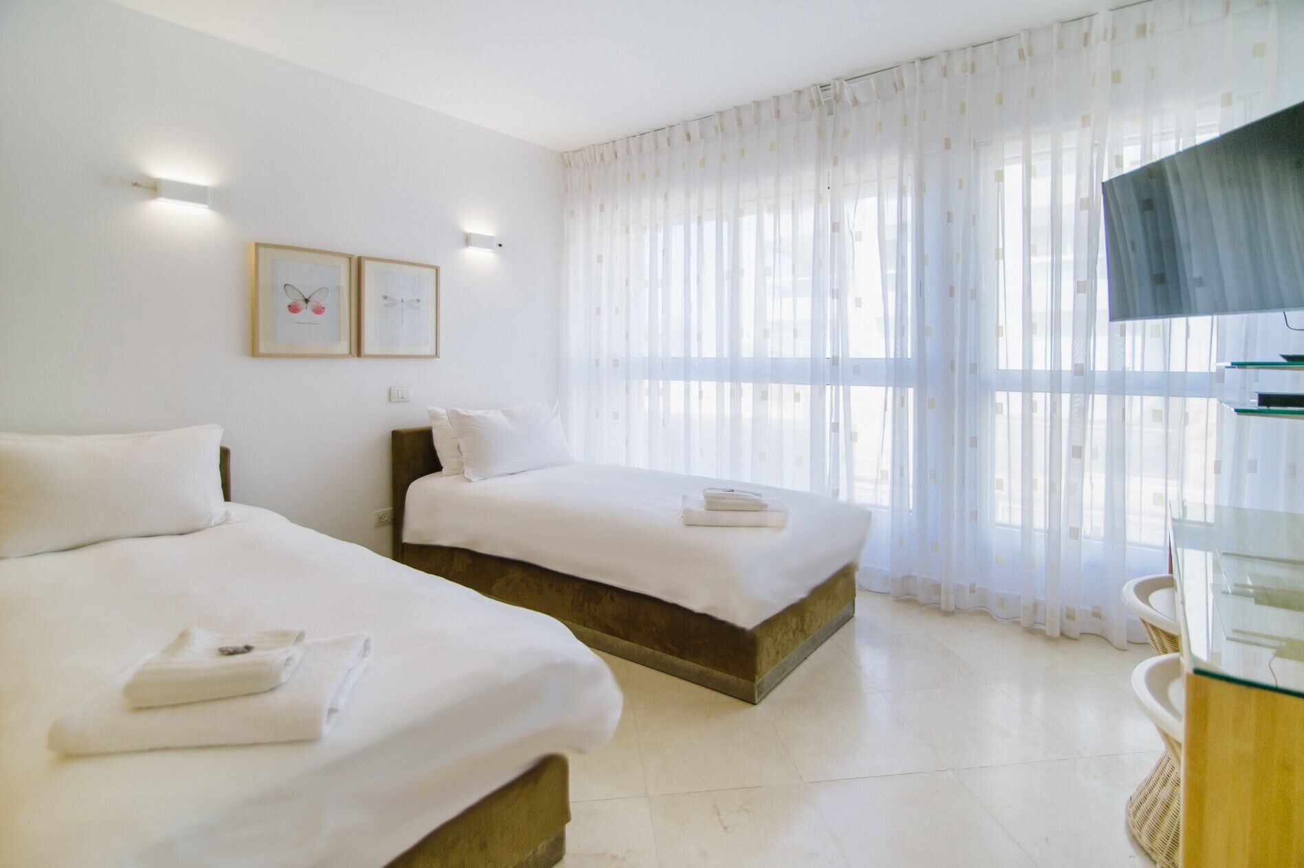 A hotel room with two beds, a large window with sheer curtains, a wall-mounted TV, and framed artwork.