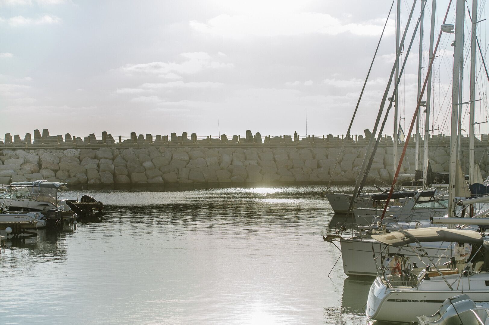 Un puerto deportivo con barcos amarrados, un muro de piedra al fondo y agua tranquila que refleja las embarcaciones.