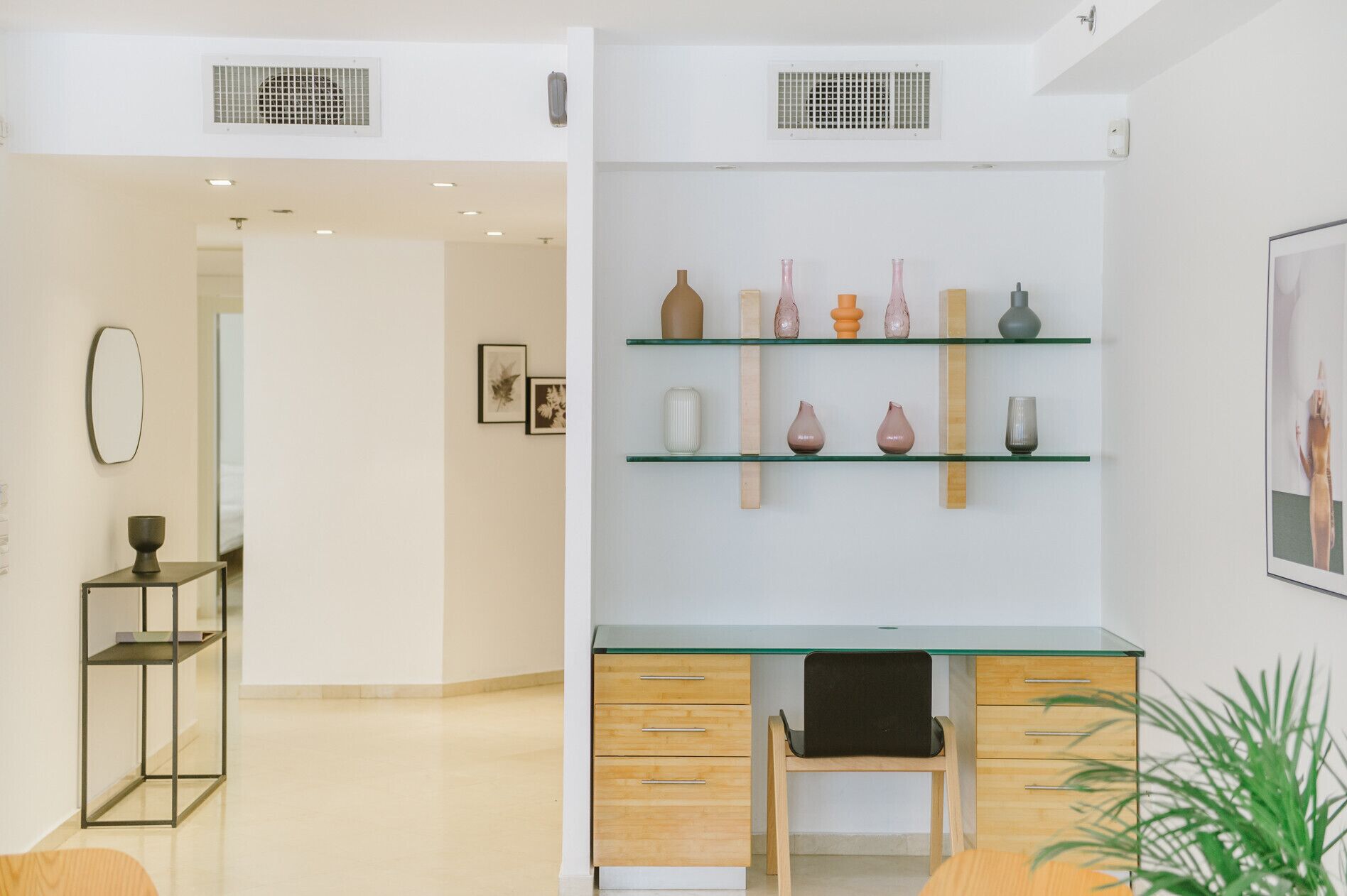 A modern office space with a glass desk, wooden drawers, and a chair. There are shelves with decorative vases and framed pictures on the wall.