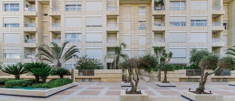 A multi-story residential building with balconies, a courtyard with palm trees, and a tiled pavement.