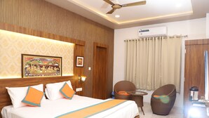 A hotel room with a bed, a ceiling fan, a wall-mounted air conditioner, a wicker chair, and a small table. - Hotel Agnija  (Udaipur)