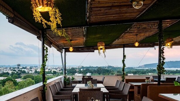 An outdoor dining area with wicker furniture, a wooden table, and hanging lights. - Hotel Agnija  (Udaipur)