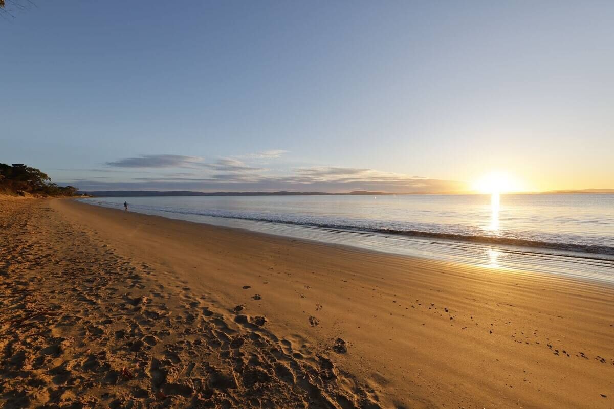 A beach with footprints leading to the water, a clear sky, and the sun setting over the horizon.