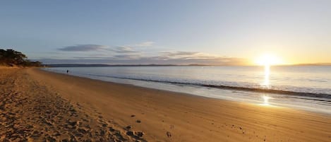 A beach with footprints leading to the water, a clear sky, and the sun setting over the horizon.