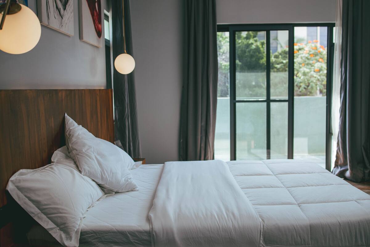 Un lit bien fait, avec du linge de lit blanc, dans une chambre dotée d’une tête de lit en bois, d’une grande fenêtre avec des rideaux et d’un luminaire suspendu.