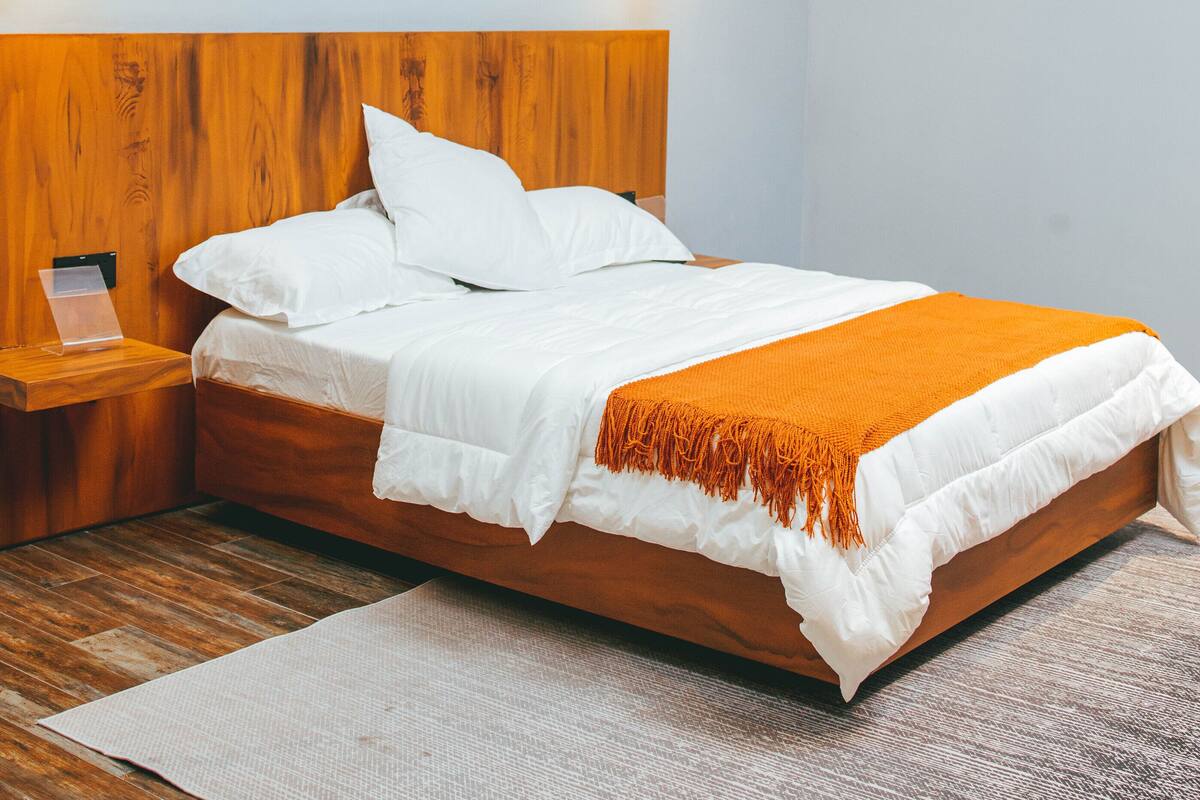 Une chambre moderne avec une tête de lit en bois, un lit avec une literie blanche et un plaid orange, une table de chevet et trois tableaux accrochés au mur.
