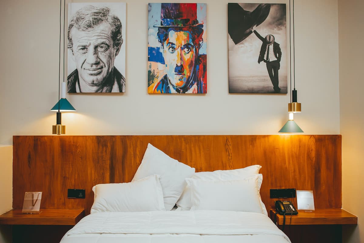 Une chambre d’hôtel avec une tête de lit en bois, du linge de lit blanc et des œuvres d’art accrochées au mur. On y trouve deux tables de chevet, chacune équipée d’une lampe, et un téléphone posé sur l’une d’elles.