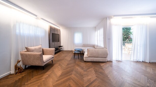 A spacious living room with a herringbone wood floor, a flat-screen TV mounted on a wall, and two beige sofas.