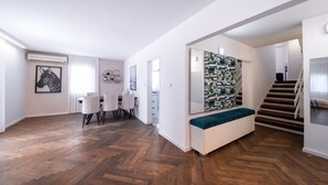 A modern living room with a herringbone wood floor, a dining area with a white table and chairs, a large abstract artwork on the wall, and a staircase leading to another level.