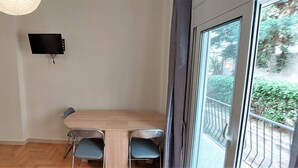 Ein Zimmer mit Holztisch und zwei Stühlen, einem flachbildfernseher an der Wand und einem großen Fenster mit Blick ins Grüne.