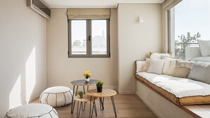 Ein kleiner, heller Raum mit einem großen Fenster, einem Sofa, zwei runden Tischen und einer kleinen Pflanze.
