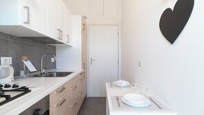 Cozinha moderna com armários brancos, pia de aço inoxidável e fogão a gás. Há uma pequena área de refeições com uma mesa posta para dois, uma taça de vinho e uma vela.