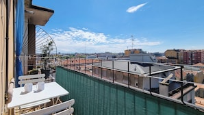 A rooftop terrace with a table and chairs, a green safety barrier, and a view of the cityscape.