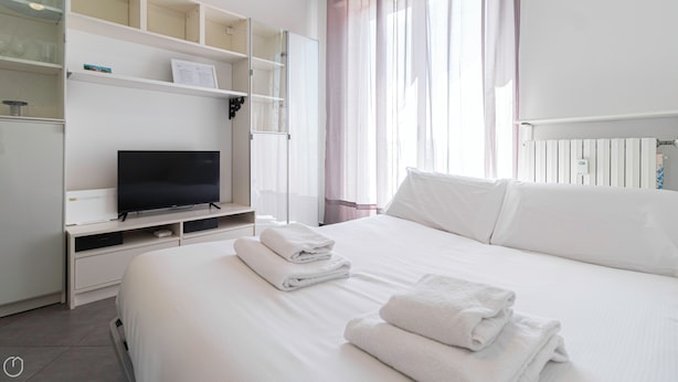 A hotel room with a bed, a TV on a stand, a shelf with a framed picture, and a window with curtains.