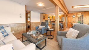 Una sala de estar acogedora con sofá, sillón y mesa de centro, vigas de madera y una cocina al fondo.