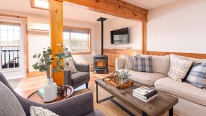 Un salón acogedor con sofá, sillón y una mesa de centro de madera. Se aprecia una chimenea al fondo y un televisor montado en la pared.