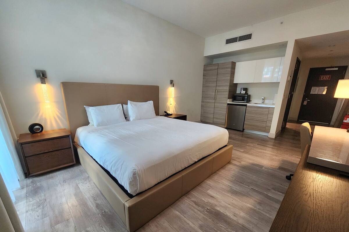 Een moderne hotelkamer met een groot bed, nachtkastjes, een ingebouwd bureau en een kitchenette.