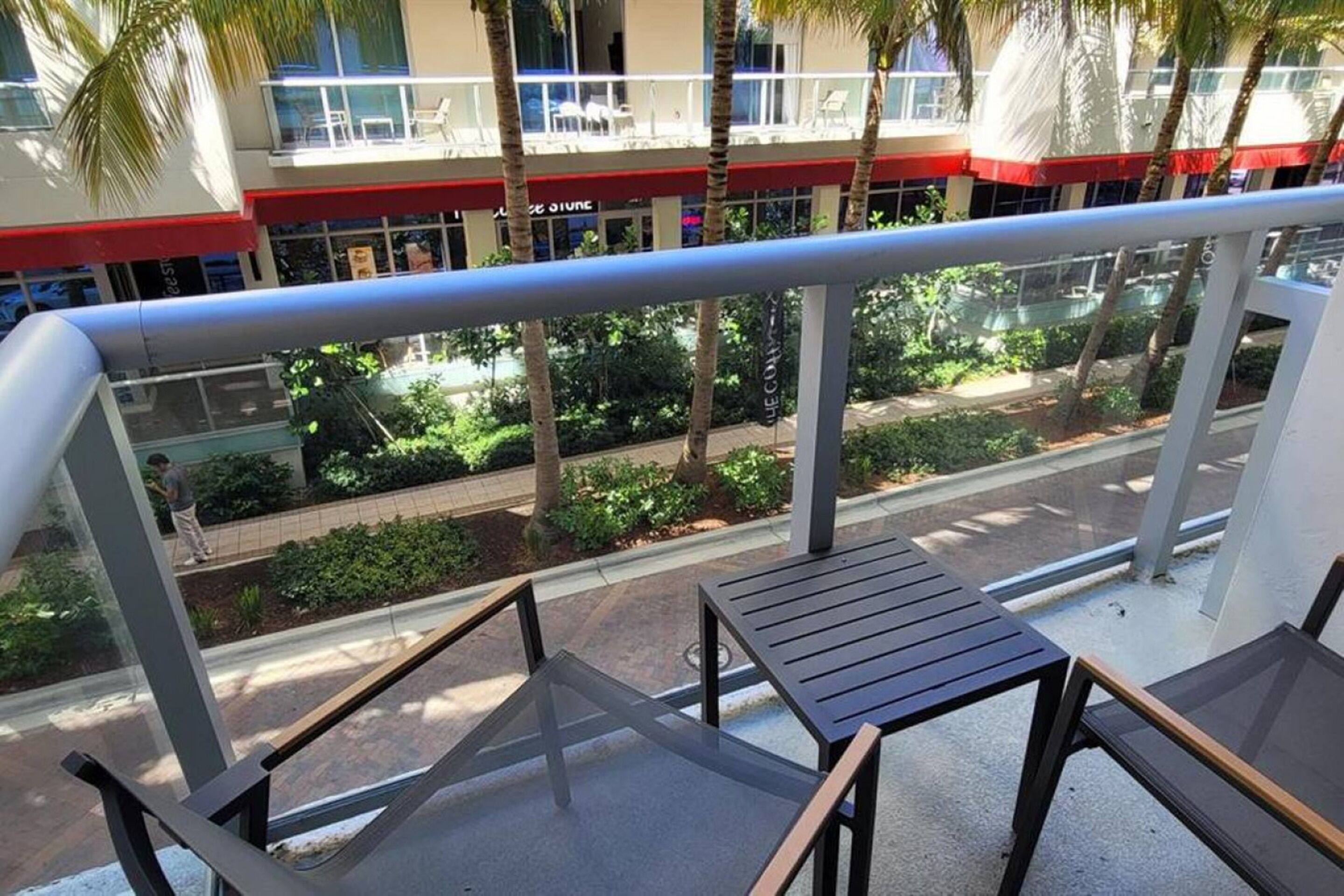 Un balcon doté d’une balustrade en verre, donnant sur une rue bordée de palmiers et sur un immeuble avec un auvent rouge.