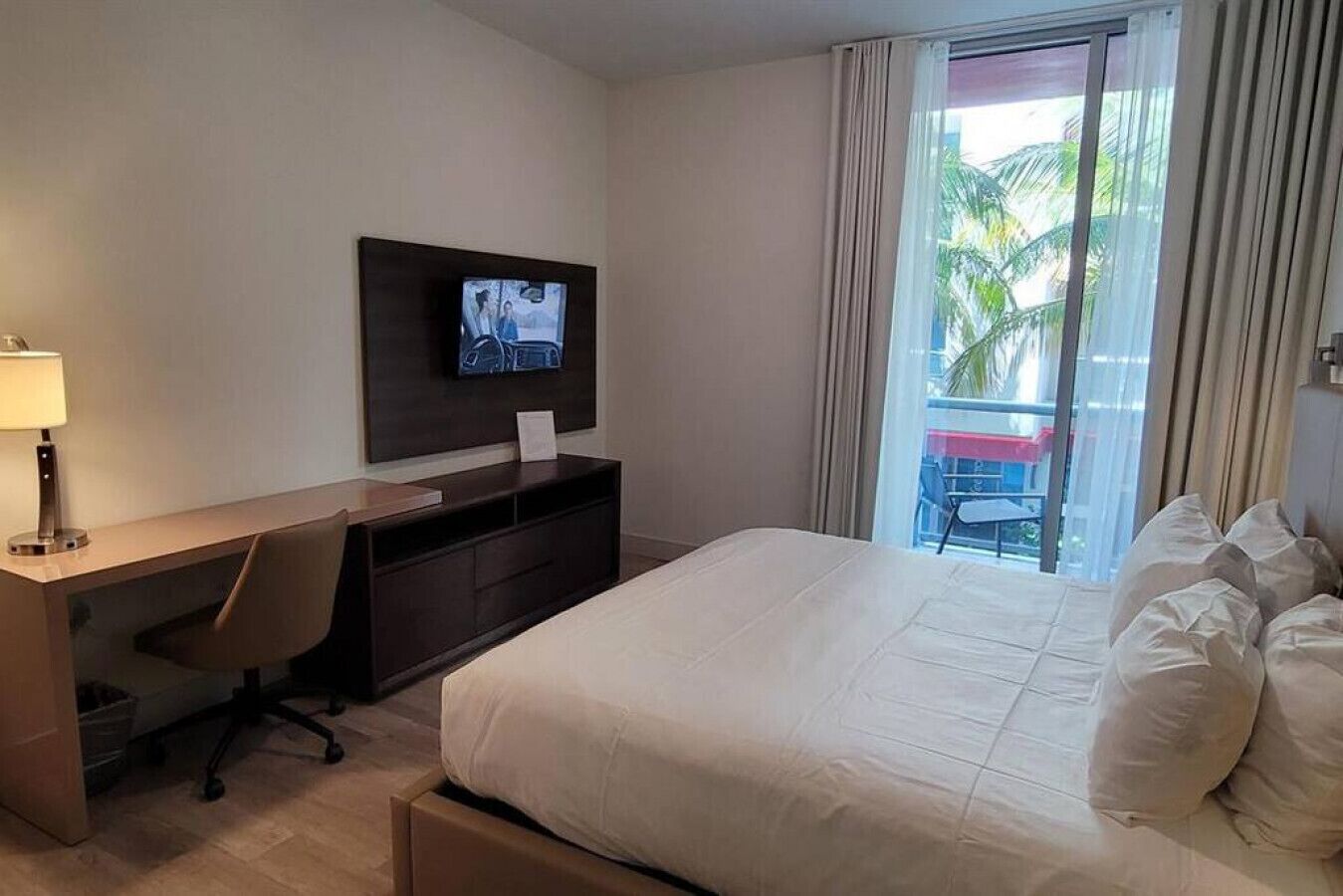 A hotel room with a bed, a desk with a chair, a TV mounted on the wall, and a view of palm trees through the window.