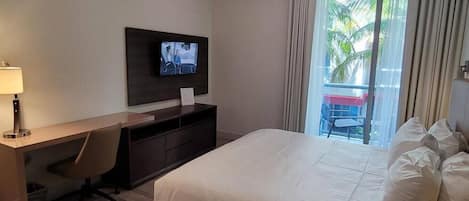 Ein Hotelzimmer mit Bett, Schreibtisch mit Stuhl, einem an der Wand montierten Fernseher und Blick auf Palmen durch das Fenster.