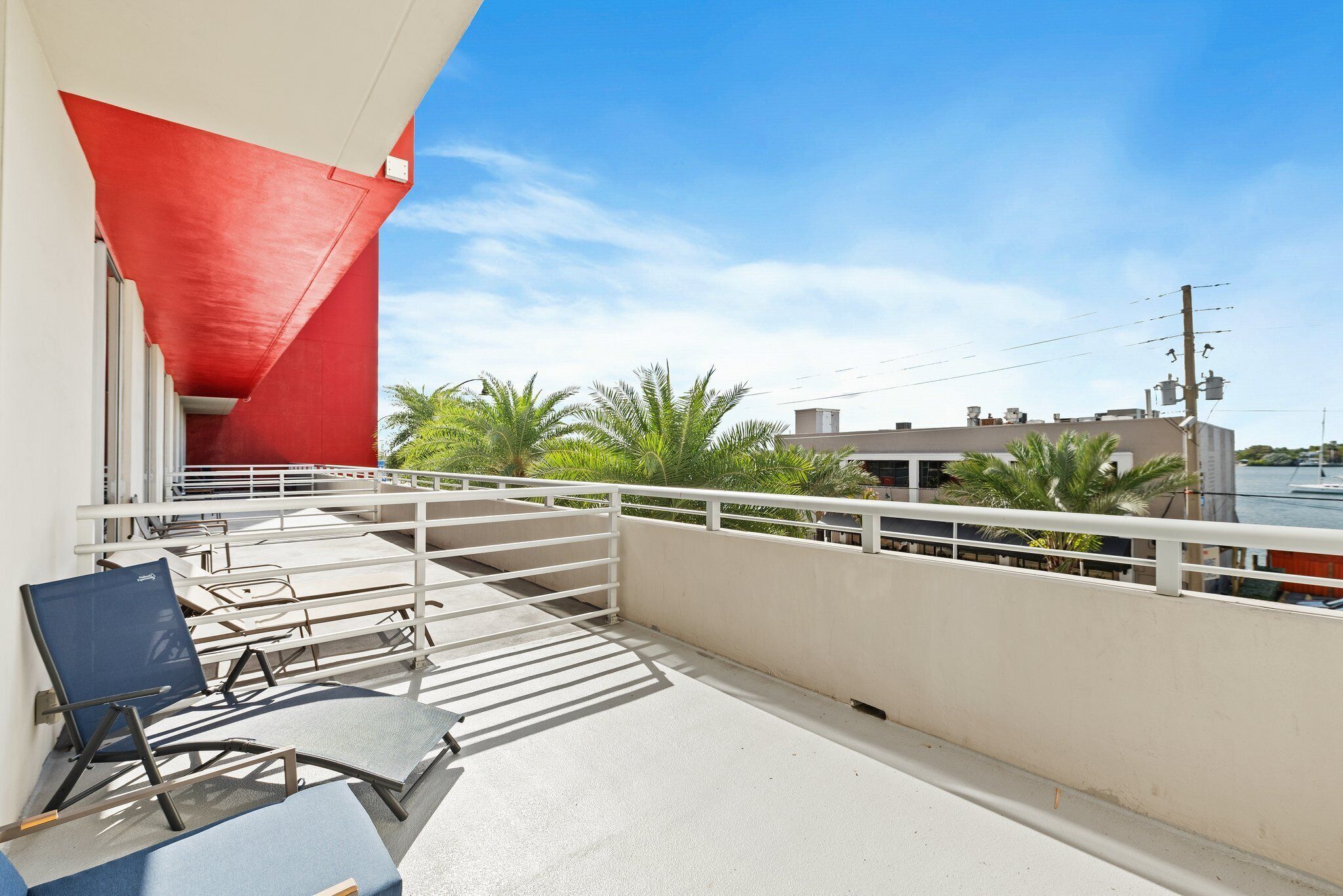 Un balcón con un elemento estructural rojo, barandilla blanca y vista a palmeras y edificios al fondo.