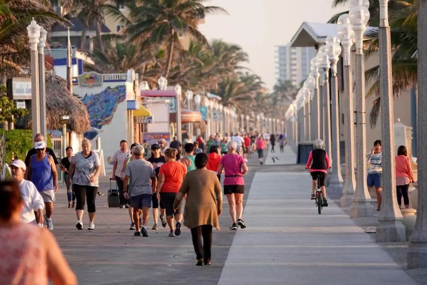 A busy pedestrian walkway lined with palm trees and buildings, with people walking and cycling.