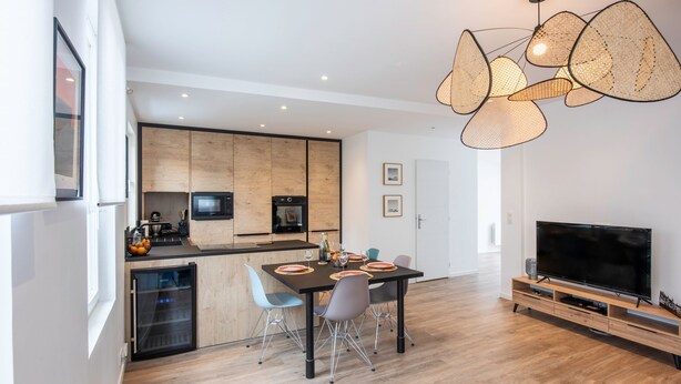 Een moderne keuken met houten kastjes, een inbouwoven en een koelkast. Er is een eethoek met een tafel en stoelen, en een flatscreen televisie op een houten standaard.