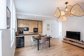 Een moderne keuken met houten kastjes, een inbouwoven en een koelkast. Er is een eethoek met een tafel en stoelen, en een flatscreen televisie op een houten standaard.
