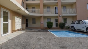 Un edifici de diversos pisos amb una zona d'aparcament, un cotxe blanc i una plaça d'aparcament reservada per persones amb discapacitat.
