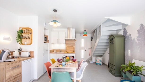 A modern kitchen with white cabinets, a wooden countertop, and a dining area with colorful chairs. - Bibifoc Baie - au coeur de la Baie de la Somme (Saint-Valery-sur-Somme)