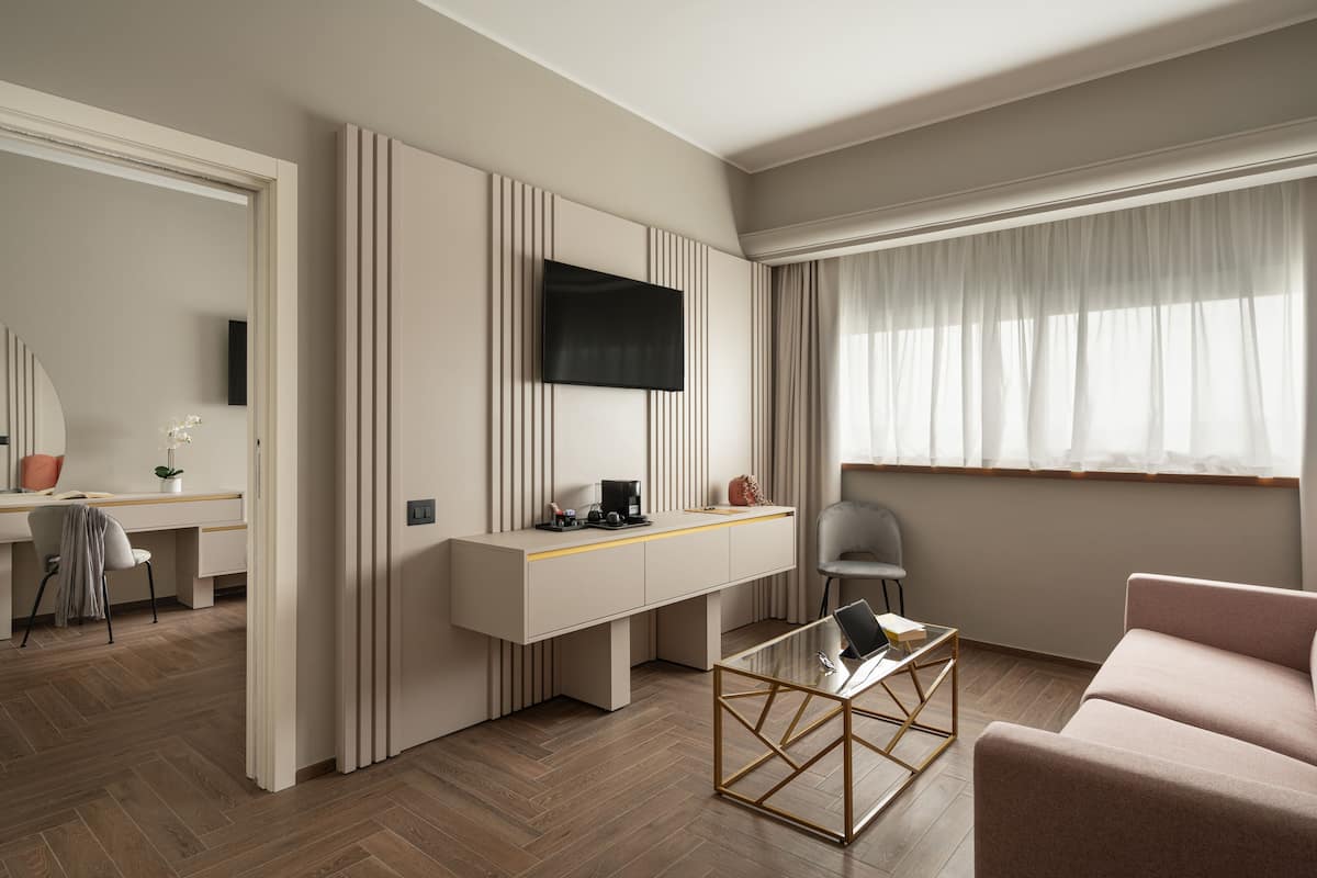 Una habitación de hotel moderna con un escritorio, un televisor montado en la pared, un sofá y un amplio ventanal con cortinas translúcidas.