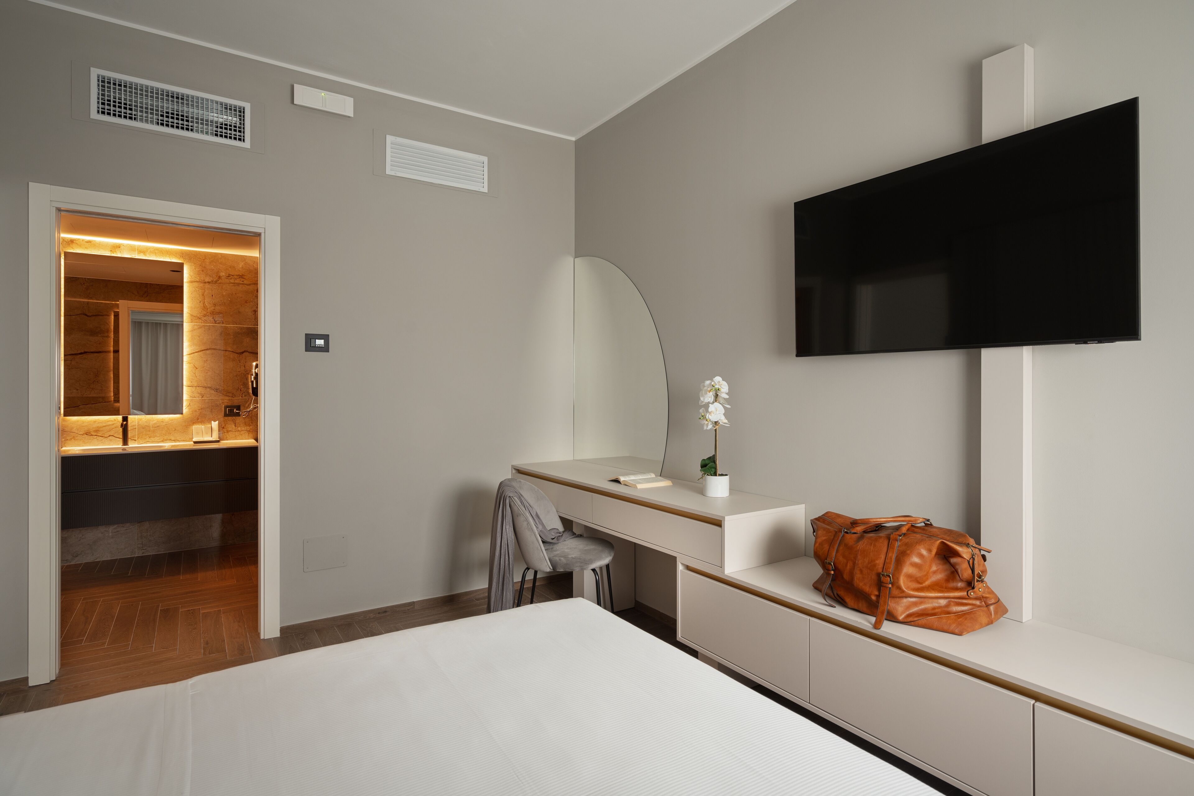 A modern hotel room with a bed, a desk with a chair, a television mounted on the wall, and a bathroom visible through an open door.