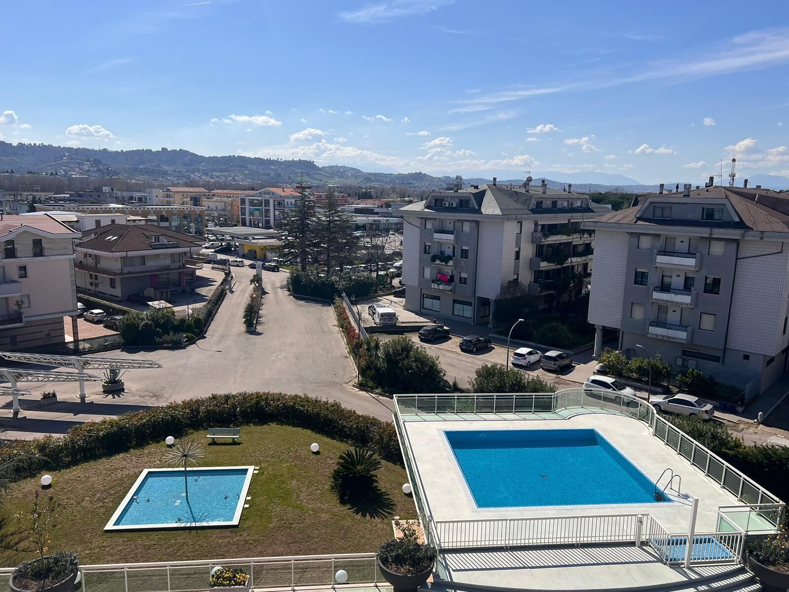 A residential area with modern apartment buildings, a central road, and a swimming pool.