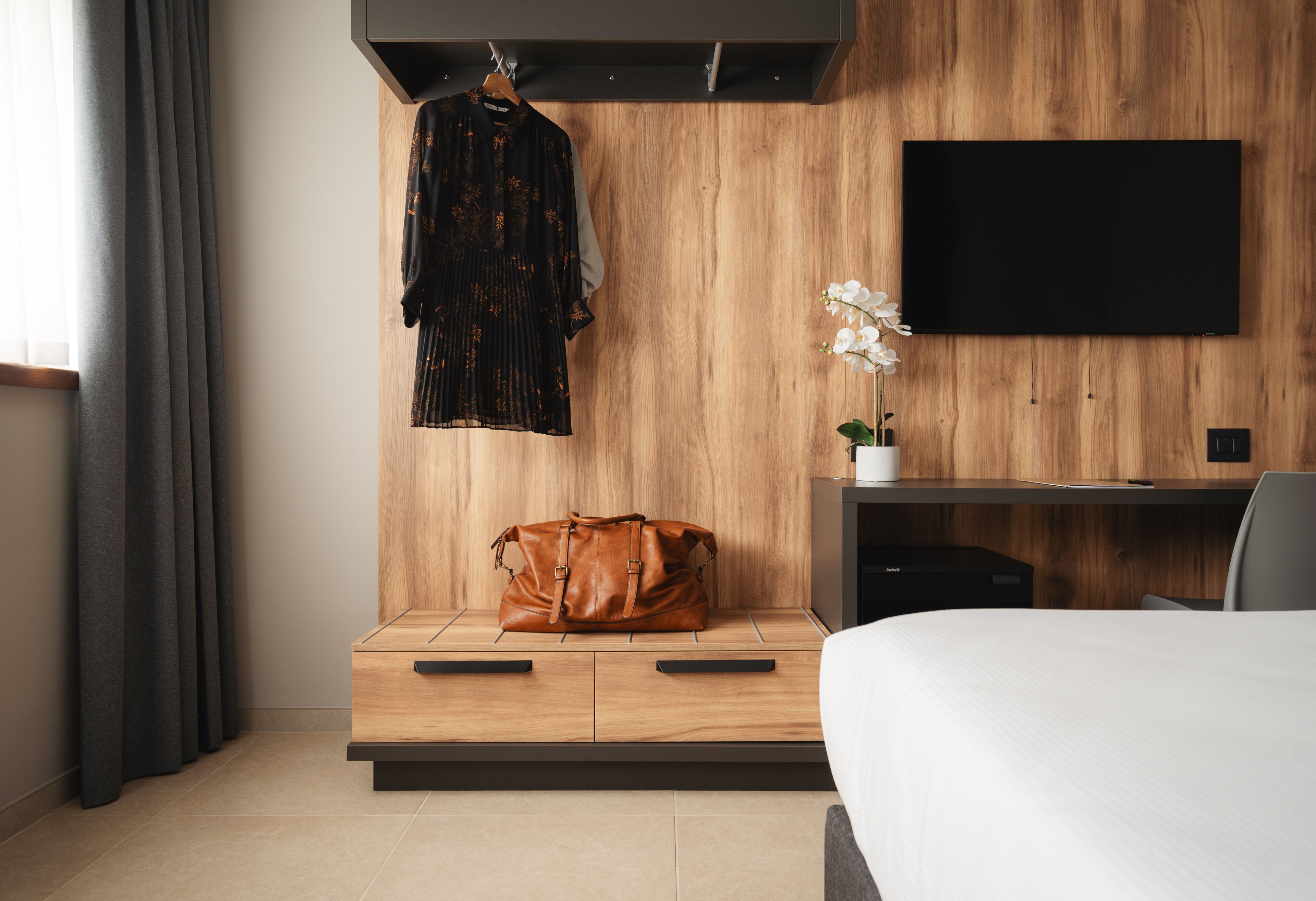 A modern hotel room with a wooden wall, a flat-screen TV, a bed, a desk, a chair, a hanging robe, a handbag, and a potted orchid.