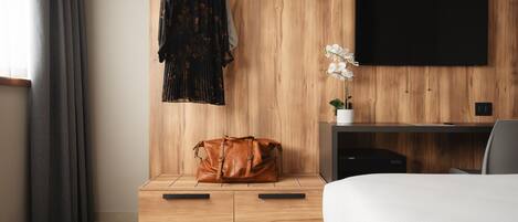 A modern hotel room with a wooden wall, a flat-screen TV, a bed, a desk, a chair, a hanging robe, a handbag, and a potted orchid.