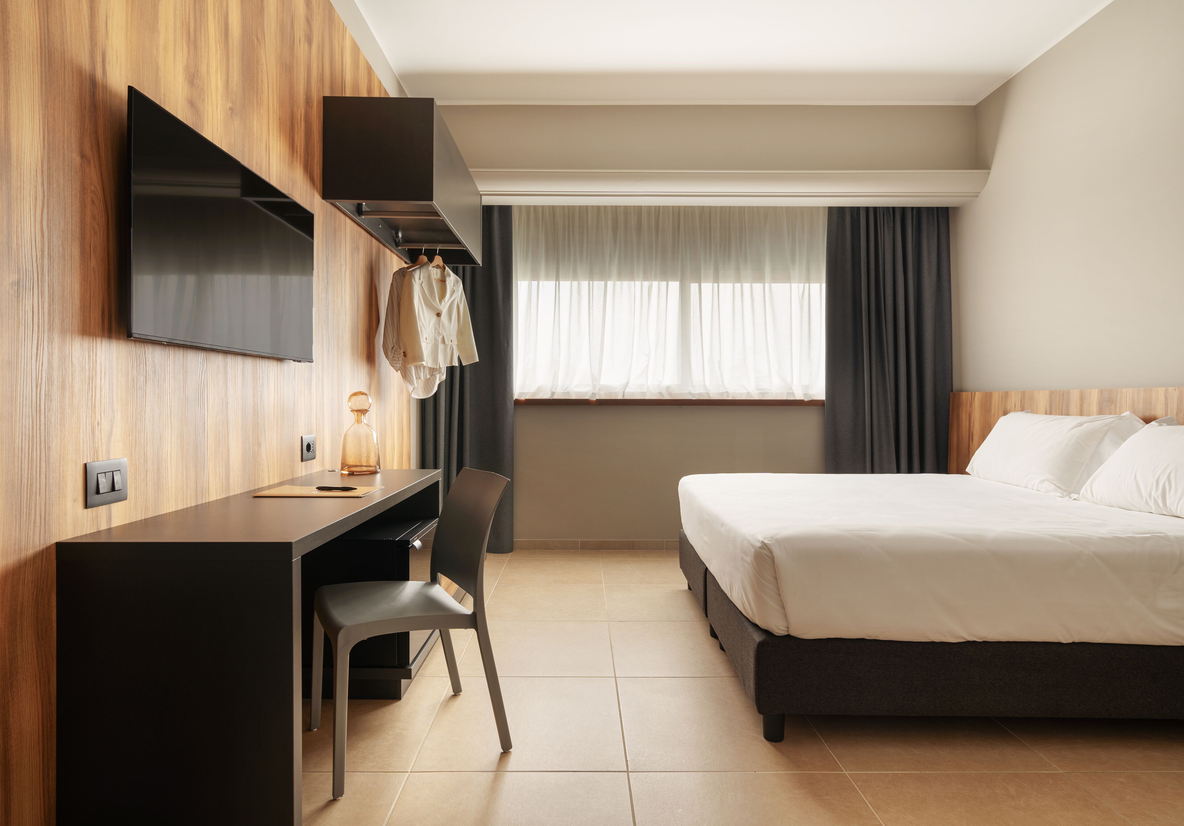 Habitación de hotel con cama, escritorio, silla, televisor montado en la pared y ventana con cortinas.