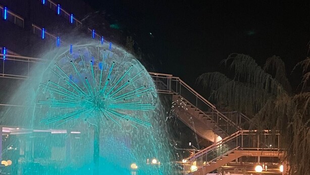 Una fuente con una estructura central iluminada por luces azules, rodeada por una piscina y una escalera que sube a un edificio con barandilla.