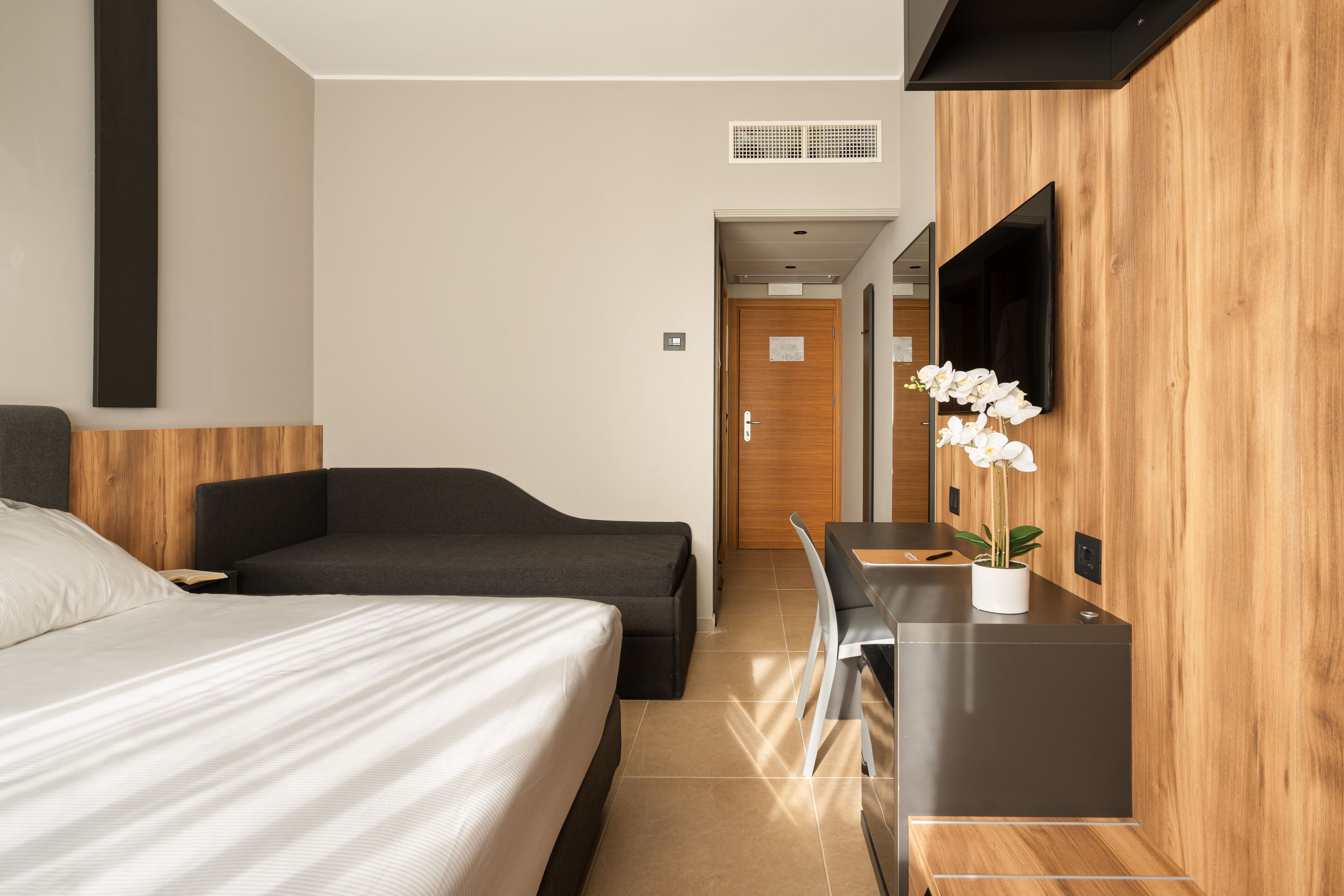 A modern hotel room with a bed, a desk, a chair, a TV, and a potted orchid.