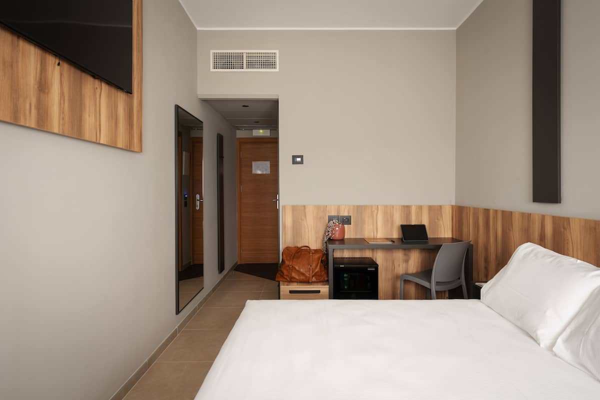 Una habitación de hotel con una cama, un escritorio con una computadora portátil, una silla, una mesita pequeña, un bolso y una puerta que da a otra habitación.