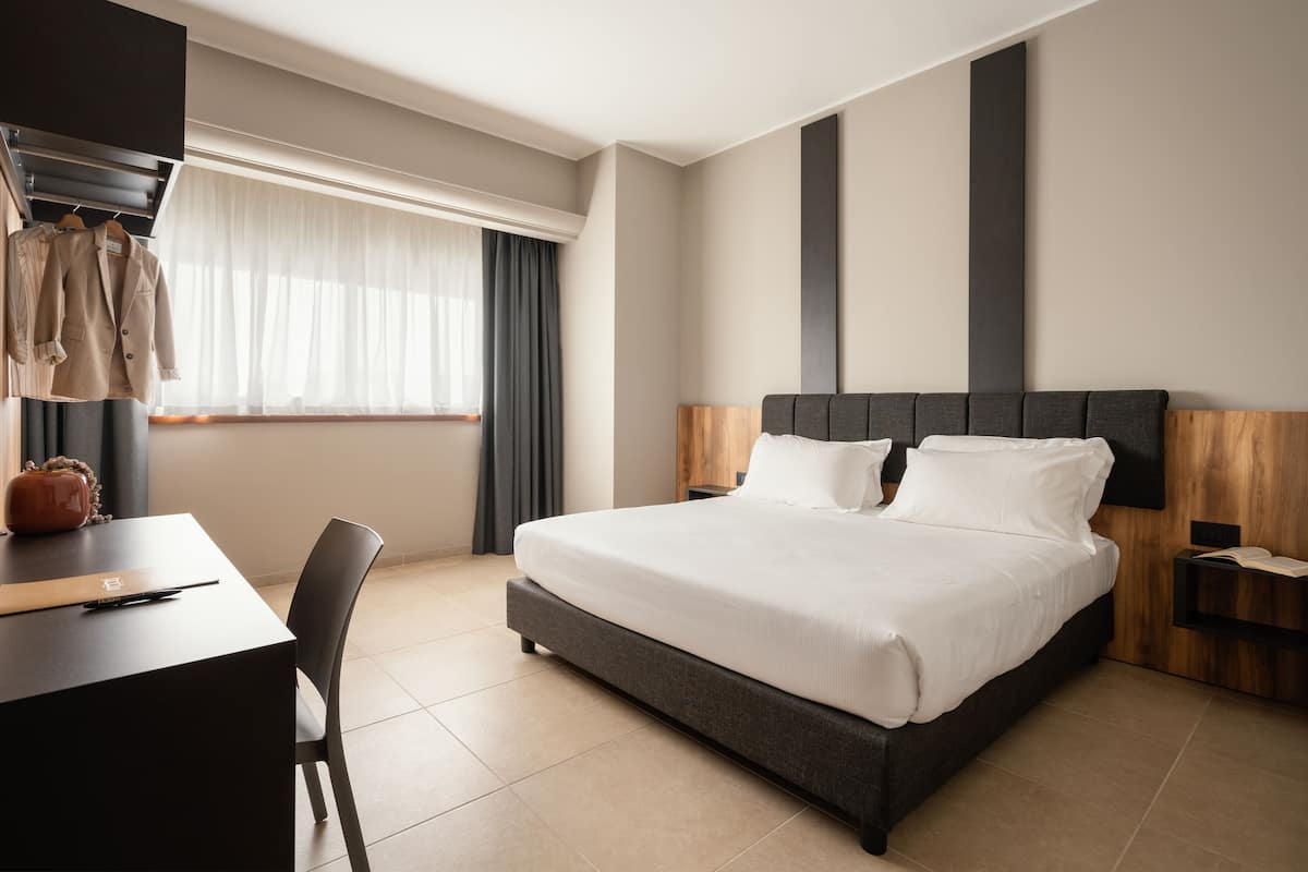Una habitación de hotel moderna con una cama grande, un escritorio con silla, un armario y una ventana con cortinas.