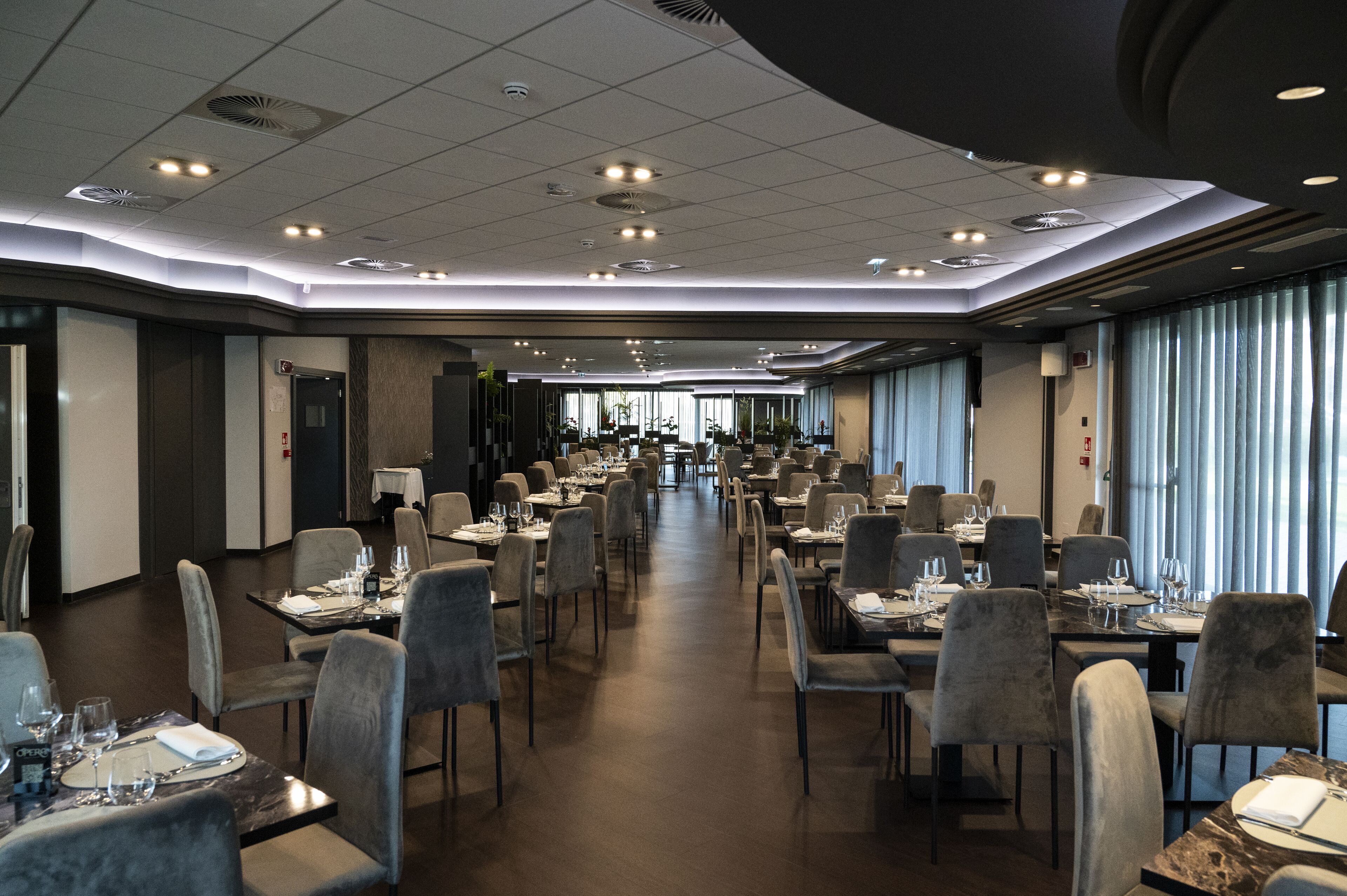 A spacious hotel dining area with multiple tables and chairs, a central walkway, and large windows with curtains.
