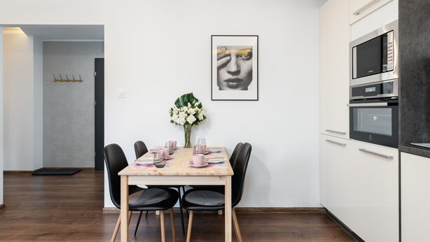 Een moderne keuken met witte kasten, een ingebouwde oven en een magnetron. Er staat een eettafel met stoelen en een bloemetjesvaas.
