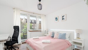 Een slaapkamer met een bed, een bureau, een stoel, een raam met gordijnen en een uitzicht op gebouwen.