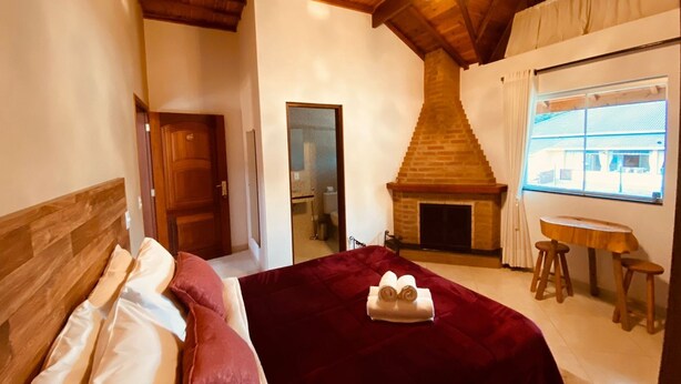 A bedroom with a wooden ceiling, a fireplace, a bed with a red blanket, and a window with a view. - Davilas Inn (Camanducaia)