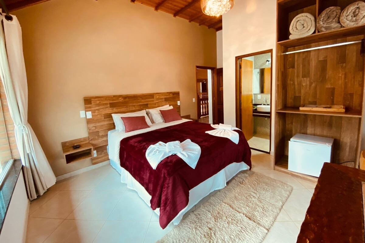 A bedroom with a wooden headboard, a large bed with a red blanket, a fluffy rug, and a wooden shelf with folded towels.