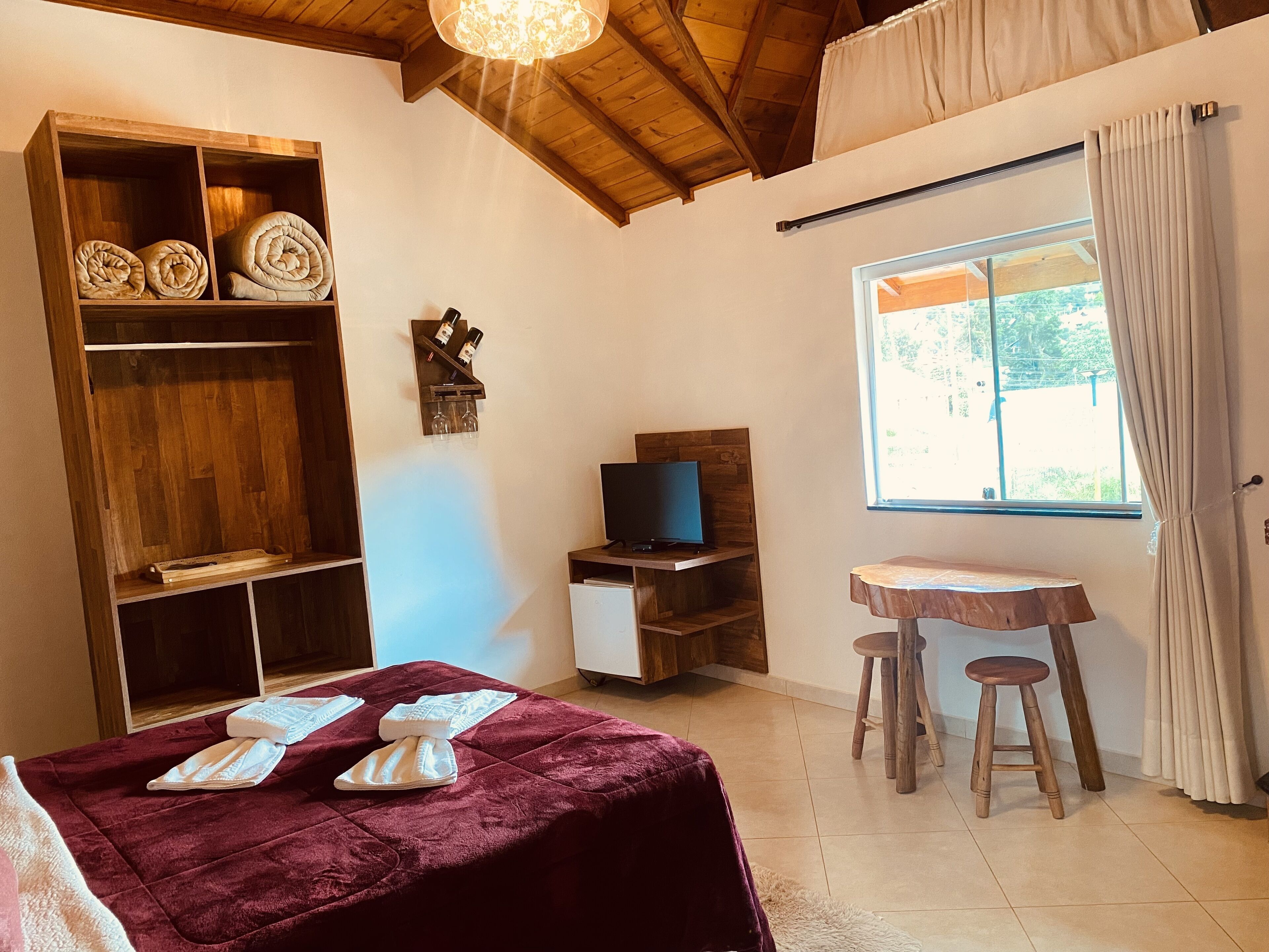 Une pièce avec une bibliothèque en bois, un téléviseur posé sur un meuble en bois, une petite table en bois avec deux tabourets et une fenêtre avec des rideaux.