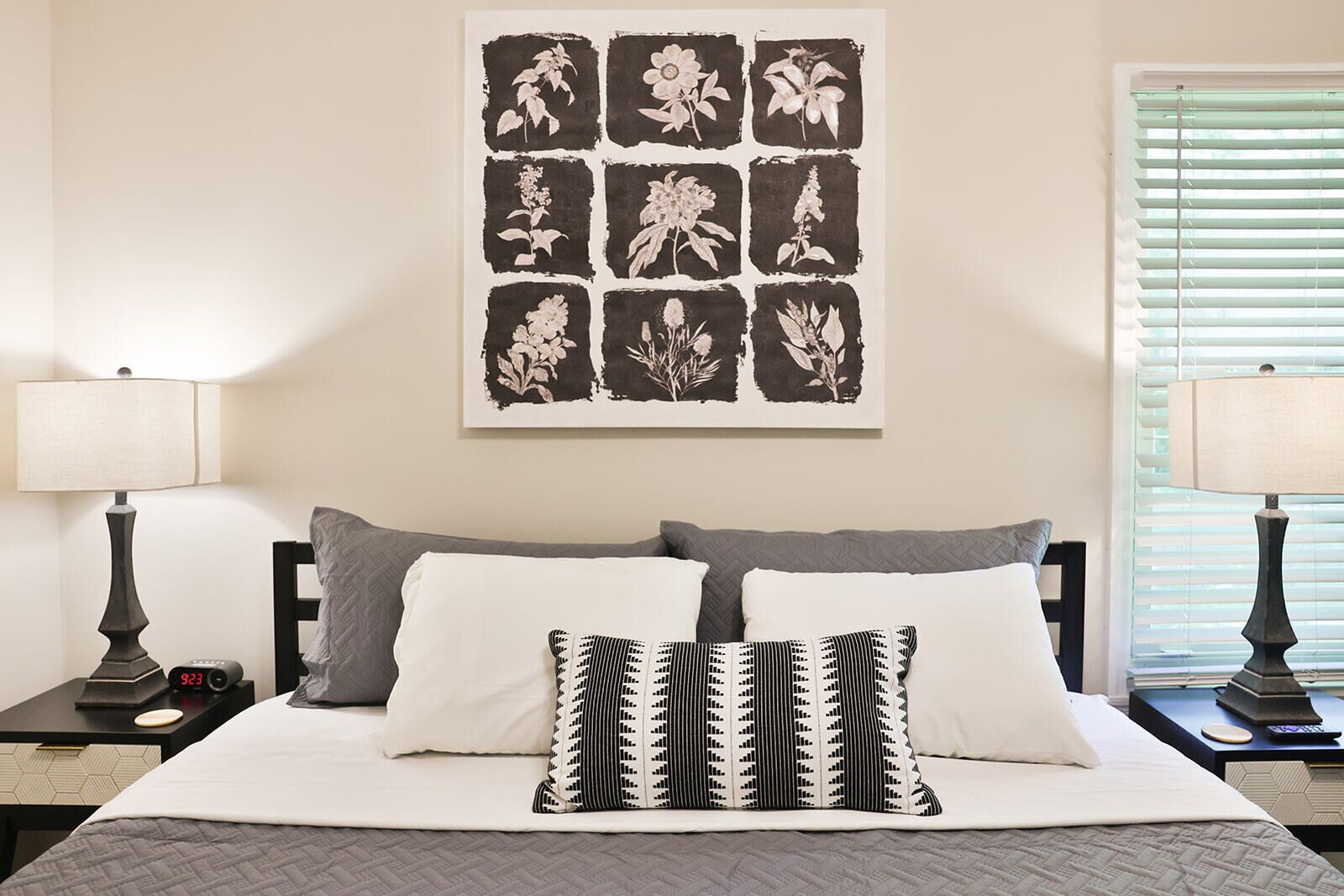 A neatly arranged bedroom with a bed, two bedside tables, a wall-mounted artwork of floral patterns, and a window with blinds.